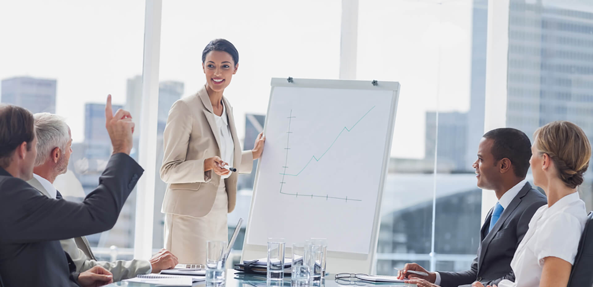 image of people in a boardroom, dressed in professional attire, looking at someone standing at the head of the table with a whiteboard drawing a graph.