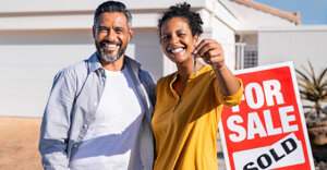 two individuals in front of a house with a "sold" sign holding keys.