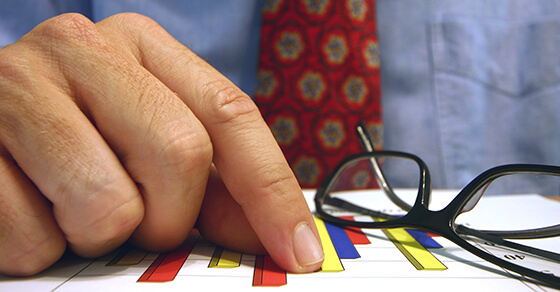 image of a piece of paper on a table with a graph on it. Next to the paper is a pair of reading glasses and a finger is pointing to a line on the chart.