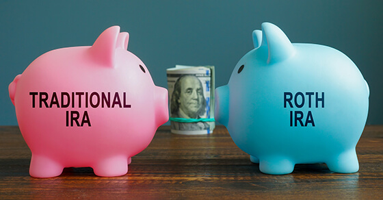 image of two piggy banks on a desk. One is pink and the other is blue. The pink piggy bank has "traditional IRA" and the blue piggy bank has "ROTH IRA" on it. In the center of the two piggy banks is rolled money.