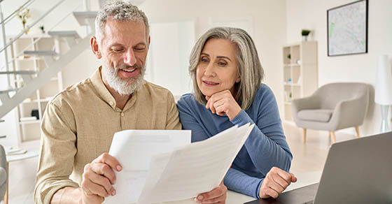 Middle aged senior old couple holding documents reading paper bills paying bank loan online, calculating pension fees, payments, taxes, planning family retirement money finances using laptop at home.