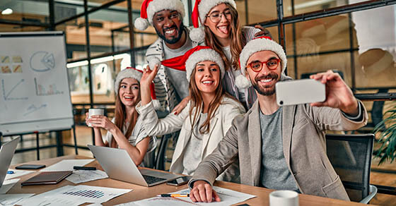 Happy New Year! Multiracial young creative people in Santa hats celebrate a holiday in a modern office and take a selfie with a smartphone on their last working day.