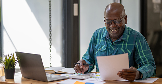 A senior man in casual clothes is busy at work but happy with the work and paperwork in front of him.